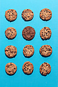 Flat lay photography of twelve chocolate chip cookies arranged on a vibrant blue background. One cookie is different from the others creating an artistic and appetizing display