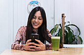 Positive female sitting at table with smoothie and browsing mobile phone while chilling at weekend in bar