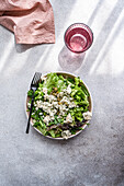 A nutritious herb salad featuring a mix of lettuce, green onion, parsley, tarragon, fermented capers, and crumbled blue brie cheese in a ceramic bowl, accompanied by a glass of water