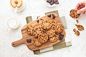 A delightful display of freshly baked chocolate chip cookies arranged on a wooden cutting board with dark chocolate pieces and a cup of milk on the side A hand picks up a cookie, suggesting a cozy, home-baked scene