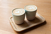 Close-up of two ceramic cups filled with latte art coffee, served on a wooden tray, placed on a similarly toned wooden table.