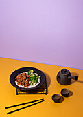 Artfully plated teriyaki chicken on a black plate, served with broccoli and rice, accompanied by a traditional tea set on a vibrant yellow and purple background.