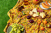 A vibrant summer picnic scene showcases a floral yellow blanket laid on lush green grass, decorated with an assortment of fruits, cheeses, and snacks