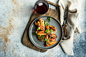 Top view roasted chicken wings in a spicy sauce garnished with fresh coriander, presented on a plate alongside a glass of cherry cocktail, with cutlery on a rustic wooden board.