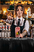 Young waitress preparing a cocktail by pouring alcohol into a jigger from the bottle in a night pub