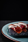 A well-composed photograph captures thinly sliced serrano ham arranged elegantly on a dark slate serving board, placed on a teal ceramic plate The rich colors and delicate texture of the ham highlight its quality and appeal