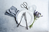 A tasteful summer table setting featuring a white plate, silver cutlery, a glass, and a bouquet of fragrant lavender on a textured background