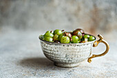 Fresh, organic gooseberries in an ornate, vintage silver bowl, showcasing their vibrant green and purple hues on a subtly textured grey background.