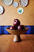 A tasteful display of shiny red apples in a rustic bowl on a wooden table, with decorative ceramic plates adorning the wall in the background.