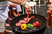 Cropped unrecognizable person grilling sausages and vegetables on a barbecue at an outdoor gathering. Background includes guests socializing, enjoying drinks and music.