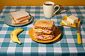 Ein hausgemachtes Frühstück auf einer karierten blauen Tischdecke mit einem Sandwich, einer Banane, Kaffee und Butter vor gelbem Hintergrund