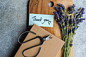 A rustic-themed thank you note beside a bouquet of fresh lavender flowers, with scissors and a wrapped gift on a wooden cutting board