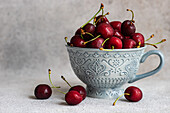 Decorative grey ceramic mug filled with juicy red cherries. The cherries overflow onto a textured grey surface, highlighting their vibrant color and fresh appearance.