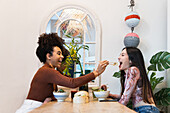 Side view of cheerful black woman feeding female friend with poke while chilling in restaurant together