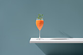 An exquisite cocktail with a fresh green sprig sits atop a white ledge against a serene blue backdrop, casting a delicate shadow.