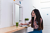 Smiling female sitting at counter with green smoothie in bar writing in notepad while speaking on smartphone