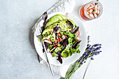 A vibrant bowl of vegetable salad with fresh lettuce, olives, juicy tomatoes, crisp cucumber, radishes, and red basil herb, sprinkled with both white and black sesame seeds, served alongside a refreshing iced drink and decorative lavender sprigs.