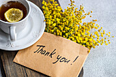 Top view of delicate cup of tea with a slice of lemon beside a cheerful bouquet of yellow Mimosa flowers and a heartfelt Thank You note