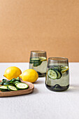 Two glasses of refreshing cucumber and lemon infused water sit beside fresh lemons and cucumber slices on a wooden tray, against a neutral backdrop.