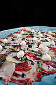 A fine dining dish of Serrano ham carpaccio garnished with shaved Parmesan cheese, fresh herbs, and dollops of cream, presented elegantly on a blue textured plate.