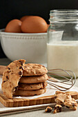 A stack of chocolate chip cookies on a wooden board, with eggs and a jar of milk in the soft-focus background, ready for baking