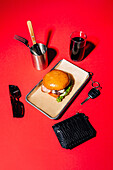 An artistically arranged barbecue meal featuring a savory burger on a metal tray, accessorized with a knife, fork, and a glass of dark beverage, contrasted against a deeply red backdrop Additional items like sunglasses and wallet