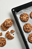 Freshly baked chocolate chip cookies cooling on a black baking tray lined with parchment paper, some broken, showcasing a tempting treat