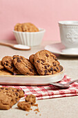 A ceramic plate overflowing with chocolate chip cookies, a spoon of sugar, and a ruffled cup on a pink backdrop create a homey vibe