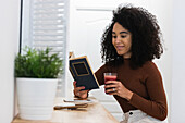 Side view of smiling black female sitting at counter in bar while reading book and drinking refreshing cocktail