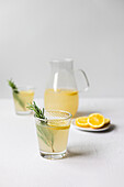 Fresh homemade lemonade with rosemary, served in a glass with a pitcher in the background on a white surface