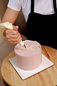 A baker decorates a pink cake with white icing, writing a message in cursive script for a special occasion