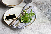 A charming table setup for summer featuring white ceramic plates accented with fresh mint leaves, vintage silverware, and a small chalkboard tag, all arranged on a gray linen napkin against a textured surface