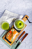 Top view of a compelling food arrangement showcasing a balanced meal that includes a green apple, cucumber and apple juice, smoked salmon on wholegrain bread, and a cherry tomato, all neatly presented on a striking marble surface.