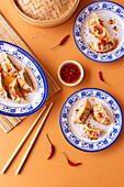 A top-down view of a steamed dumplings garnished with chili flakes and served with a spicy chili dipping sauce, elegantly presented on blue and white decorative plates, accompanied by a bamboo steamer, chopsticks, and a bowl of chili sauce, all set against an orange background