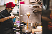 A chef wearing a red turban is intently preparing food in a busy commercial kitchen, showcasing culinary expertise with focus and authenticity