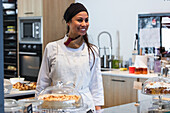 Positive female baker in uniform standing at counter with vegan desserts in backhouse and looking away