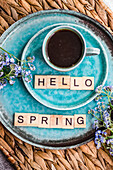 A cup of coffee and forget-me-not flowers on a turquoise plate with Hello Spring spelled out in letter tiles, evoking a fresh springtime mood