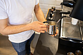 A barista in a casual attire steams milk with a professional espresso machine, focusing on the shiny metal pitcher.