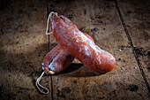 A pair of traditional sobrasada sausages, a specialty from the Balearic Islands, laid on an aged wooden table. The sausages are rich in color, indicative of their paprika content.