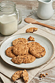 A cozy arrangement of chocolate chip cookies on a ceramic plate, paired with a glass jar of milk and a wooden spoon, all portrayed on a rustic wooden table