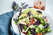 Colorful mixed vegetable salad featuring lettuce, ripe tomatoes, crunchy cucumbers, sliced radishes, black olives, and avocado, sprinkled with black and white sesame seeds and red basil leaves. Served in a white bowl with a fork on the side, perfect for a healthy meal.