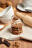 Stack of chocolate chip cookies bound with twine, placed next to baking tools on a wooden table, evoking a warm, homemade feel