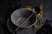 From above an artistic table setting featuring sleek black dinnerware, accented by vibrant yellow mimosa flowers and a touch of greenery.