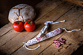 A rustic wooden table displays traditional Spanish fuet, sliced cured sausage, fresh tomatoes, and crusty bread, showcasing a blend of Mediterranean flavors.