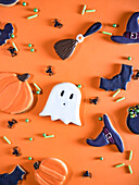 A festive array of Halloween-themed cookies including shapes of ghosts, witch hats, and bats, creatively arranged on a bright orange background, complimented by candy sprinkles and decorative spiders