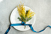 From above of ornate white plate holds a bright bouquet of mimosa flowers, elegantly tied with a deep blue ribbon on a textured grey surface