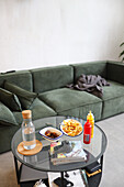 A cozy home setup showing a relaxing day on a green modern sofa, enjoying snacks, consisting of French fries and dates, with a neutral decor backdrop.