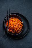 Freshly grated carrot salad served in a matte black bowl with a pair of black chopsticks, presented on a dark textured background