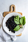 Top view of a white plate full of juicy, dark mulberries garnished with fresh green leaves, presented on a rustic wooden board. The image is composed to give a sense of natural and organic freshness, perfect for conveying health and vitality.