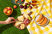 Close-up of a vibrant summer picnic spread on a checkered yellow blanket over green grass, featuring an array of fruits, cheeses, and cured meats with anonymous hand reaching to adjust a cheese slice